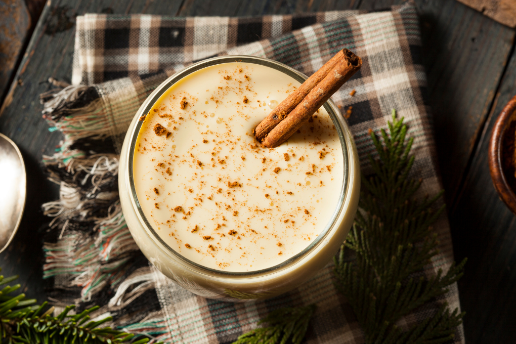 Close-up of a glass of creamy eggnog garnished with a sprinkle of nutmeg and a cinnamon stick. Blog post about eggnog and where to find it locally.